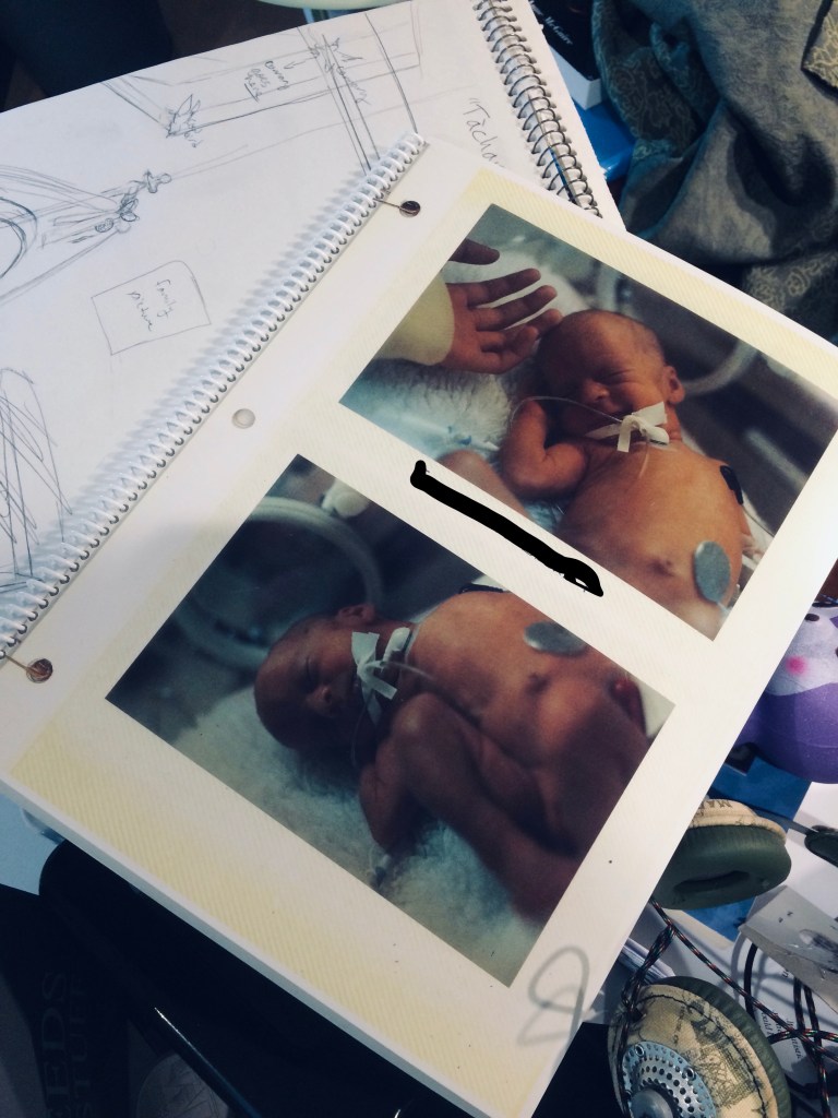 A photo album is shown resting on top of a sketchbook where the composition of a painting is being worked out. The open page has two photos of a baby in an incubator, wires and monitors are glued to its chest and stomach, and an IV tube is taped just below its mouth. In the upper photo the infant is leaning into a hand that is extended from the left of the frame.