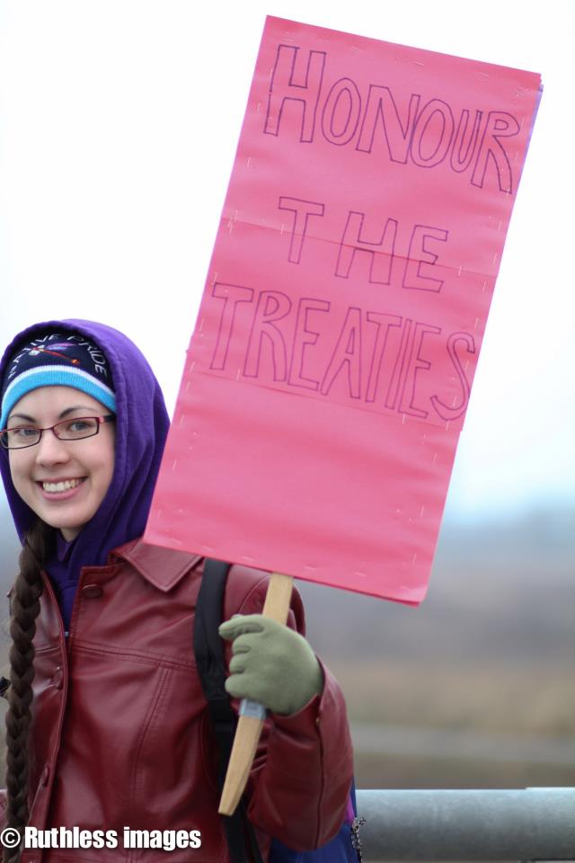 An image of the author holding a sign saying "Honour the Treaties". They are wearing a red jacket and a hat that says "Native Pride".